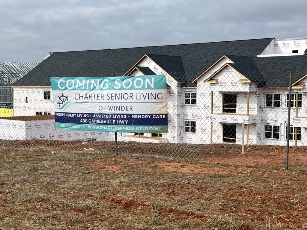 banner on fence that reads, "Coming Soon, Charter Senior Living of Winder, Independent Living, Assisted Living, Memory Care, 436 Gainesville HWY" in front of community under construction